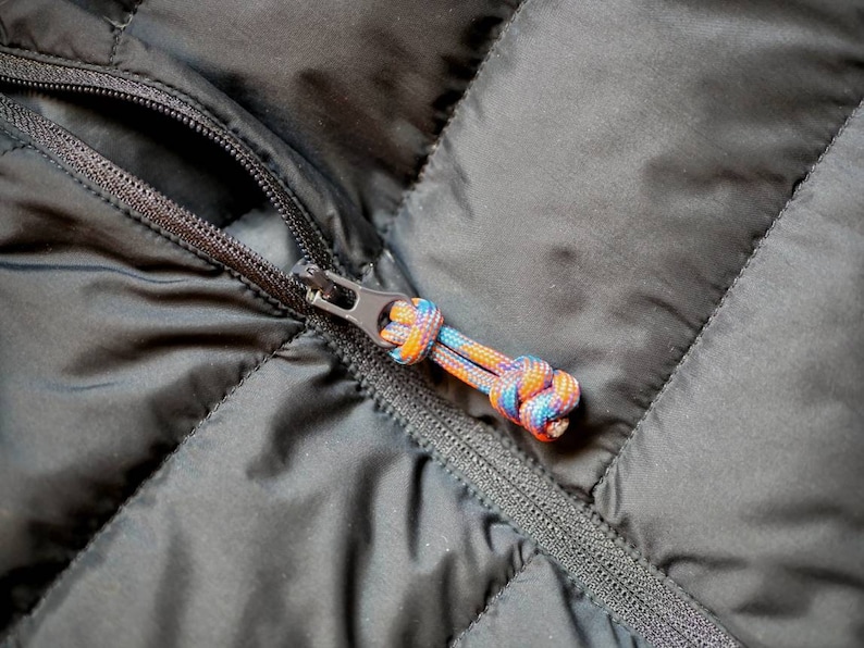 May include: Close-up of a black quilted jacket zipper with a colorful, braided pull. The zipper is closed, and the pull features a mix of blue, orange, and yellow cords. The jacket's fabric has a diamond pattern.