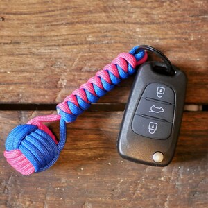 May include: A pink and blue paracord keychain with a black car key fob attached. The keychain is made of a braided rope with a knot at the end.
