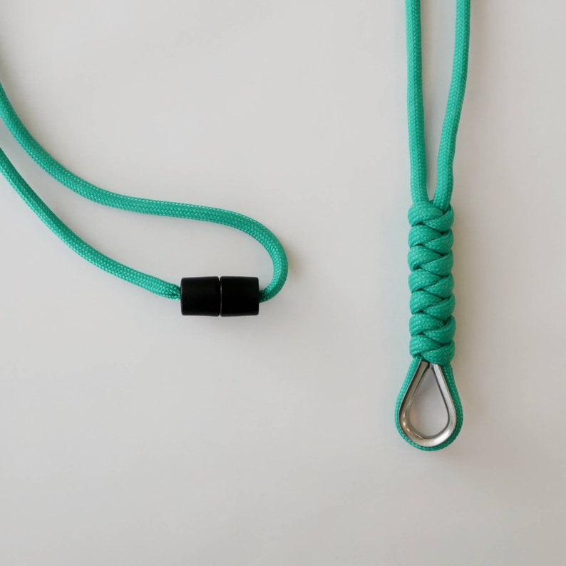 K&ouml;nnte beinhalten: Ein t&uuml;rkisfarbenes Lanyard mit einem schwarzen Kunststoff-Schieber und einem silbernen Metallhaken am Ende.
