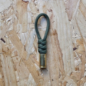 Puede incluir: Un llavero de paracord verde oscuro con una carcasa de bala de latón. El llavero está atado en un nudo y tiene un lazo en la parte superior.