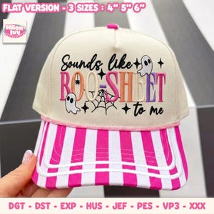 May include: A cream-coloured baseball cap with a pink and white striped brim. The front features embroidered text that reads Sounds like BOO-SHEET to me, accompanied by two small white ghost figures, a black spider, and a spider web design.