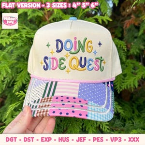 Könnte beinhalten: Eine cremefarbene Trucker-Cap mit dem gestickten Text DOING SIDE QUEST in bunten Buchstaben und kleinen Sternen. Der Schirm zeigt ein farbenfrohes Patchwork-Muster aus Streifen, Punkten und Vichy-Karo. Der Text gibt Größen von 10, 13, 15 cm an.