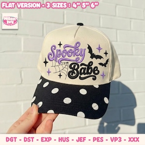 Puede incluir: Una gorra de béisbol color crema con visera negra y lunares blancos. El panel frontal muestra el texto bordado Spooky Babe en morado y negro, acompañado de una telaraña negra y dos murciélagos negros, con pequeños detalles de estrellas moradas.