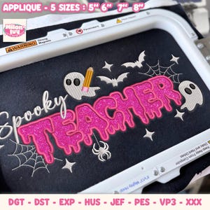 May include: An embroidery design on black fabric featuring the text Spooky Teacher in bright pink glittery font with a dripping effect. The design includes white ghosts, bats, a spider, spiderwebs, stars, and a yellow pencil. Text reads APPLIQUE - 5 SIZES : 5" 6" 7" 8".