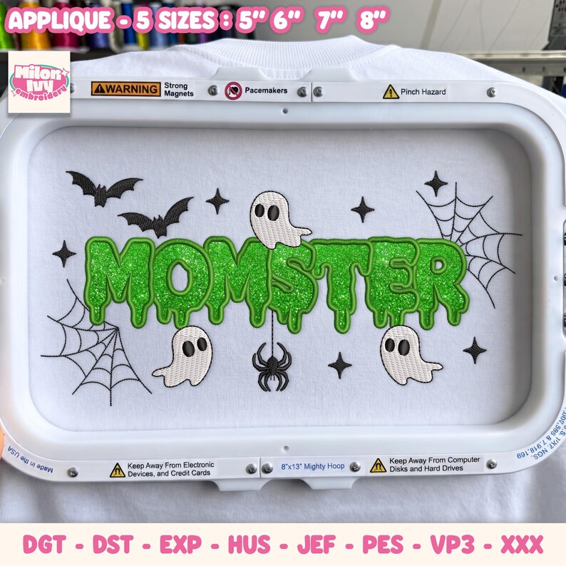 May include: A white fabric surface featuring an embroidered design with the green glittery text MOMSTER in a dripping font. The design includes black bats, white ghosts, black spiders, spider webs, and small black stars. The fabric is held in a white hoop.
