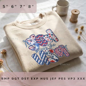 May include: A beige sweatshirt with the phrase "Bless the USA" in red, white, and blue patterned letters. The design includes floral and striped patterns, with a bow detail. The sweatshirt is folded.