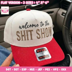 May include: A white baseball cap with a red brim and a red button on top. The front features black embroidered text that reads welcome to the and gold embroidered text that reads SHIT SHOW. A sticker on the brim displays OTTO Classic Fit.