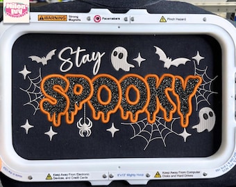 May include: An embroidery design in a white rectangular hoop featuring the text Stay SPOOKY in black glitter and orange thread. The design includes white ghosts, bats, spiderwebs, and a spider on a black fabric background. Text states APPLIQUE - 5 SIZES : 5" 6" 7" 8".