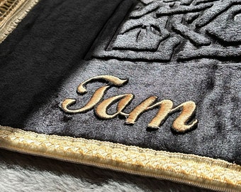 May include: A black prayer rug with a gold embroidered border and a gold embroidered design in the center. The rug has the name "Jam" embroidered in gold on the bottom.