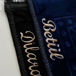 May include: Two prayer rugs, one black and one navy blue, are displayed. The black rug has the name "Diana" embroidered in gold thread, while the blue rug features "Betül". Both rugs have decorative trim and fringe.