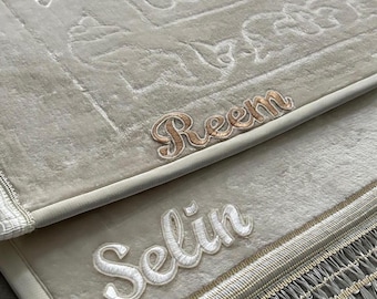 May include: Two beige prayer rugs with embroidered names, "Reem" and "Selin", in gold and white thread. The rugs have a white fringe border.