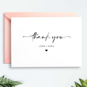 May include: A white thank you card with the words "thank you" in elegant black script. Below the text are the names "JOHN + SARA" and a small black heart. The card is paired with a light pink envelope.