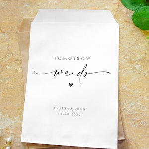May include: White wedding favor bag with the text "TOMORROW we do" in black script font with a small black heart below. The names "Caitlin & Colin" and the date "12.28.2020" are printed below in black.