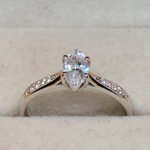 May include: A silver engagement ring featuring a pear-cut diamond. The band is set with smaller stones. The ring is displayed in a black and beige ring box, set against a yellow backdrop.