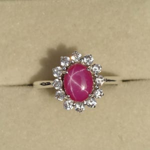 Op de afbeelding: Een zilveren ring met een ovale ster-robijn, omringd door een halo van kleine, heldere edelstenen. De ring wordt gepresenteerd in een zwarte sieradendoos met een beige interieur.