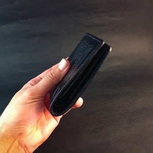May include: A black leather wallet with a textured design. The wallet is folded in half and held in a hand.