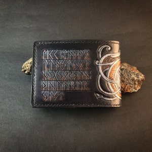 May include: Black leather wallet with a silver and brown design featuring a knotwork pattern and runes. The runes are in a grid pattern and read "M K C E F F F R T M S S X R S E X S R E I N E D N D K B M K G S T".