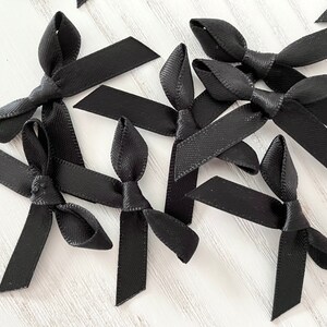 Peut inclure: Un groupe de nœuds en ruban de satin noir, noués en un simple nœud, sont disposés sur une surface en bois blanc. Les nœuds sont tous de la même taille et de la même forme, et ils sont tous noués de la même manière.