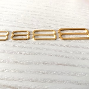 Könnte beinhalten: Vier goldfarbene Metall-Schieber-Schnallen in verschiedenen Größen. Die Schnallen sind in einer Reihe von klein nach groß angeordnet.