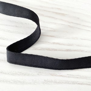 May include: Black ribbon with a slightly textured surface. The ribbon is folded in a curved shape and is lying on a white wooden surface.