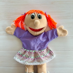 May include: A handmade puppet doll with orange yarn hair tied with purple ribbons. The doll wears a purple top and a patterned skirt with a white lace trim. The doll's shoes are gray, purple, and yellow.