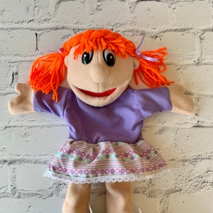 May include: A handmade puppet with a light skin tone, orange yarn hair, and a purple dress. The puppet has a smiling red mouth, black eyes, and wears purple and gray shoes with yellow accents. The skirt has a floral pattern.