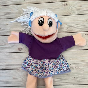 Könnte beinhalten: Eine Handpuppe mit einem lila Shirt, einem Blumenrock und weißen Haaren mit blauen Schleifen. Die Puppe trägt blaue und grüne Schuhe.