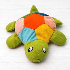 May include: A plush turtle toy featuring a soft green head and limbs, with a multi-colored shell composed of fabric patches in shades of orange, yellow, light blue, pink, and dark blue. The turtle has two black and white plastic eyes.