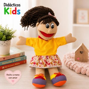 May include: A fabric hand puppet with dark brown yarn hair, a yellow shirt, and a floral skirt. The puppet has a red mouth, black and white eyes, and wears multicoloured shoes with orange, yellow, and purple panels. Text reads Didácticos Kids.