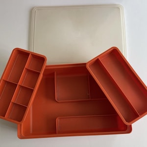 May include: An orange plastic storage tray with a matching lid. The tray has several compartments of varying sizes, including a rectangular section and a section divided into smaller compartments. The lid is a translucent off-white color.