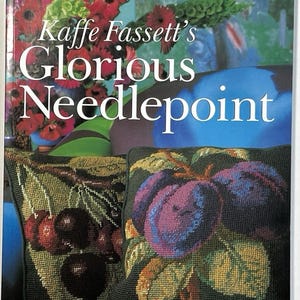 May include: A book cover titled "Kaffe Fassett's Glorious Needlepoint" with a colorful floral arrangement in the background. Two needlepoint pillows are in the foreground, one with cherries and the other with plums.