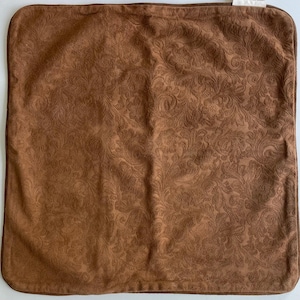 May include: A square, brown decorative pillow with a detailed, embossed floral pattern. The pillow has a darker brown border and a soft, suede-like texture. The overall design is classic and elegant.