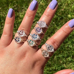 May include: A hand displays several silver wire rings, each featuring a clear, smiley face design with different colored eyes. The rings are set against a backdrop of green grass, with the hand's nails painted a lavender color.