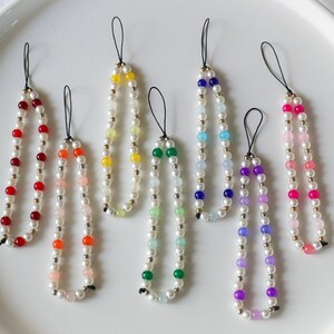Puede incluir: Una colección de siete colgantes de teléfono con cuentas. Cada colgante presenta una cadena de perlas falsas blancas, intercaladas con cuentas de colores en tonos rojos, naranjas, amarillos, verdes, azules, morados y rosas. Cuentas doradas acentúan el diseño.
