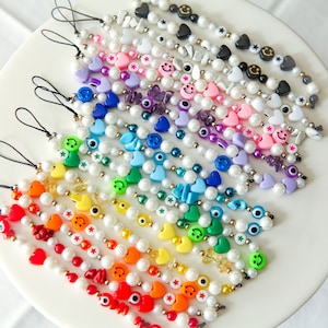 Könnte beinhalten: Eine Sammlung farbenfroher, mit Perlen besetzter Telefonanhänger. Jeder Anhänger verfügt über eine Vielzahl von Perlen, darunter Perlen, Smiley-Gesichter, Herzen und andere dekorative Elemente. Die Anhänger sind fächerförmig auf einer weißen Oberfläche angeordnet.