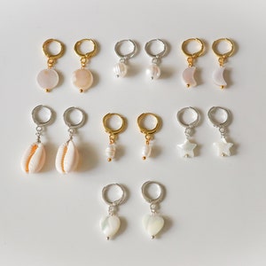 May include: A collection of earrings with various designs, including round, pearl, shell, moon, star, and heart shapes. The earrings feature gold and silver hoops. The earrings are displayed on a white surface.