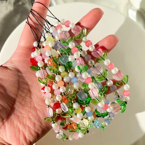 Puede incluir: Una colección de coloridos colgantes para teléfono con cuentas. Cada colgante presenta un diseño floral con cuentas rosas, verdes y transparentes, intercaladas con perlas blancas y otras cuentas de colores. Los colgantes están sujetos a cordones negros.