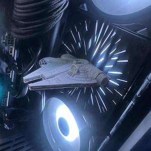May include: A detailed view inside a computer case, showcasing a miniature Millennium Falcon model. The model is positioned near a glowing white fan, with other components and wiring visible. The scene is illuminated by the fan's bright light.