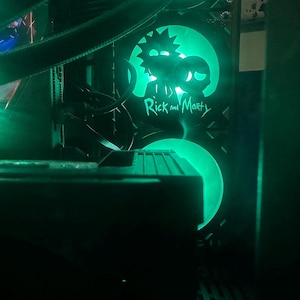 May include: A computer case with green LED lighting. The case features a design of Rick and Morty characters. The text "Rick and Morty" is visible below the character silhouettes. The overall aesthetic is dark with bright green accents.