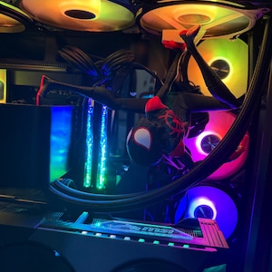 May include: A Miles Morales Spider-Man action figure hanging upside down inside a computer case. The figure is black and red with white eyes. The computer interior features glowing RGB fans in yellow, pink, and blue, along with an MSI branded graphics card component.