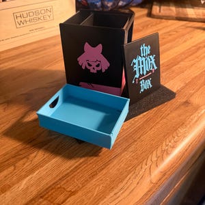 May include: A black storage container with a pink skull graphic and a detachable light blue tray. A separate black panel features the text 'the Mox Box' in a stylised blue font. The container has an open top and a divided interior compartment.