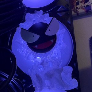Puede incluir: Una figura de personaje esférica de color negro con grandes ojos blancos y una boca roja que muestra dos colmillos blancos. La figura está rodeada por un aura translúcida, texturizada y nubosa, iluminada por una luz azul brillante desde atrás.