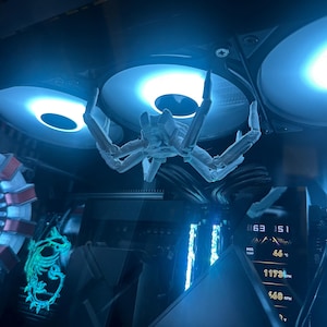 May include: A computer case interior with a white, articulated spider-like figure suspended between two illuminated cooling fans. The scene is bathed in blue light, with other internal components visible.