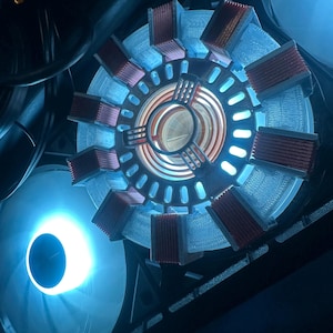 Może przedstawiać: Niestandardowy komponent komputera o okrągłym kształcie, z centralnym świecącym rdzeniem i otaczającymi prostokątnymi elementami. Urządzenie ma niebiesko-czerwoną kolorystykę, z wentylatorem w tle.