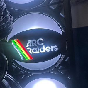May include: Close-up of a computer fan with the text "ARC Raiders" in silver lettering. The fan has a series of colorful stripes in green, yellow, and red. The fan is lit with a bright white light.