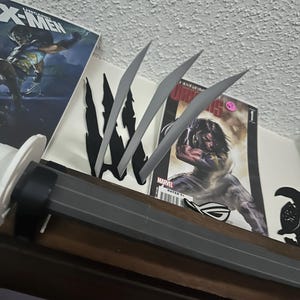 May include: A display featuring comic books and a replica of Wolverine's claws. The claws are silver and sharp, positioned above a dark grey handle. Comic books include the X-Men and Marvel series.