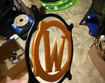May include: A circular black frame featuring a metallic orange letter W centered within a matching orange circular border. The design includes four small mounting holes positioned at the top, bottom, left, and right sides of the black outer ring.