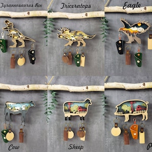 May include: Six wooden keychains with different animal designs. Each keychain has a small leather pouch and a metal ring. The animals are a Tyrannosaurus Rex, a Triceratops, an eagle, a cow, a sheep, and a pig.
