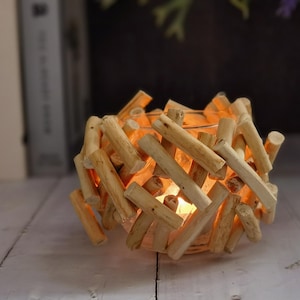 May include: A natural wood candle holder with a woven design. The candle holder is made of small, light-colored wooden sticks arranged in a crisscross pattern. A warm, soft light glows from within, creating a cozy ambiance. The candle holder sits on a white surface.