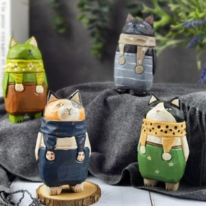 Puede incluir: Cuatro figuras de gato de madera talladas a mano. Cada gato está pintado con diferentes colores y patrones, incluyendo verde, marrón, negro y blanco. Los gatos llevan monos y bufandas. Un gato está sobre una rodaja de madera.